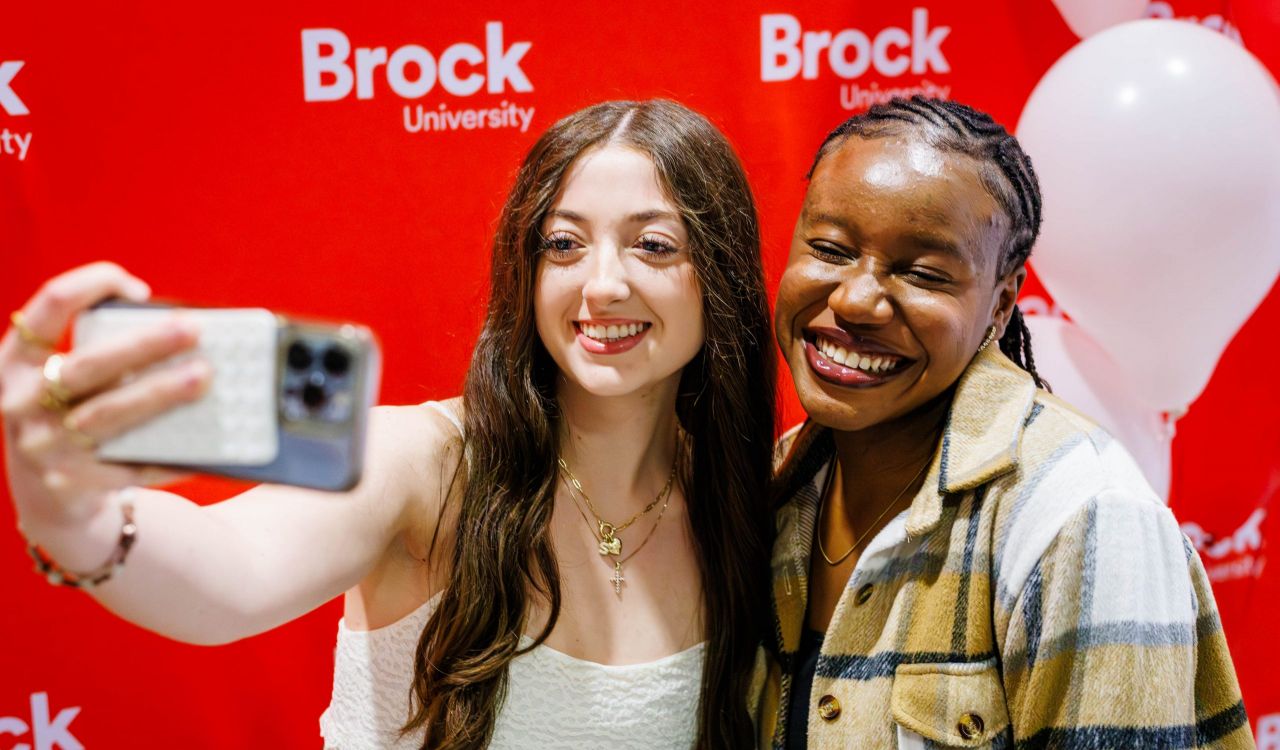 Two students take a selfie in front of a red backdrop.