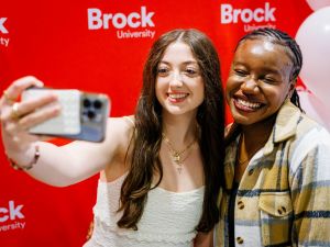 Two students take a selfie in front of a red backdrop.