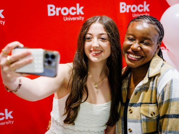 Two students take a selfie in front of a red backdrop.
