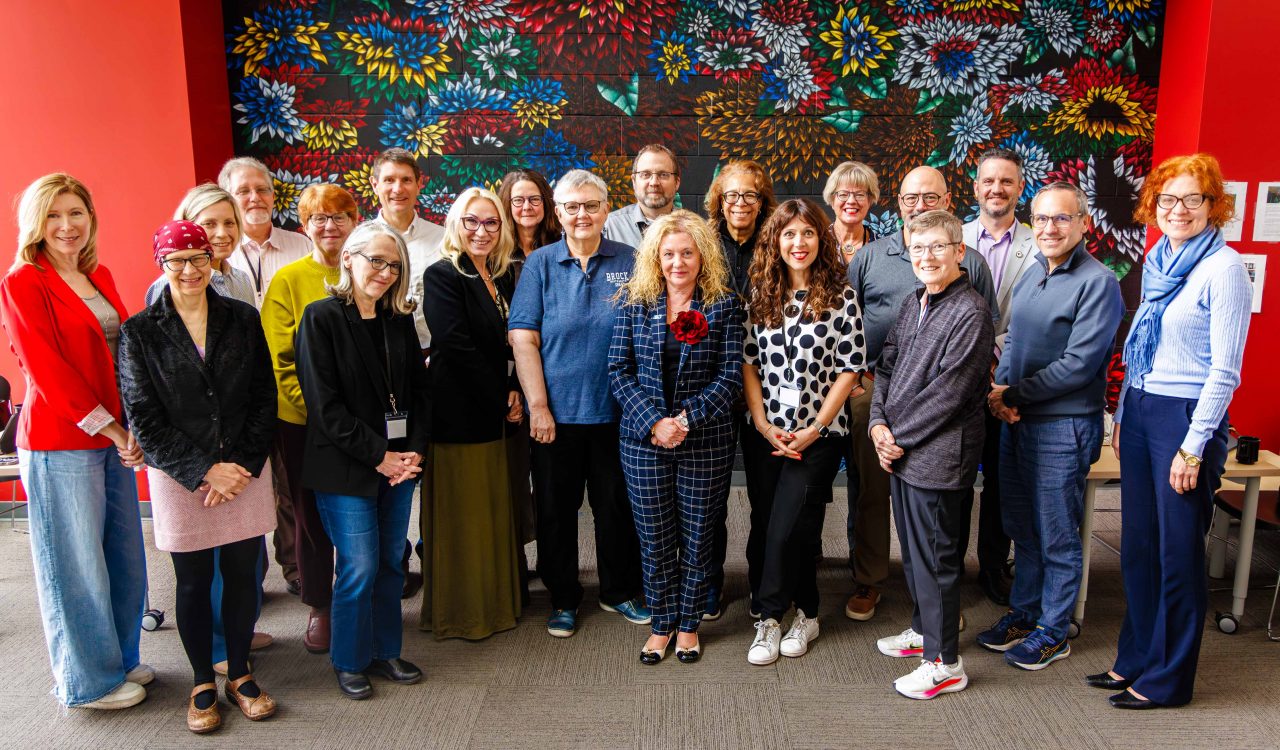 A large group of university staff pose together in front of a colourful floral mural during a Provost Summit gathering.