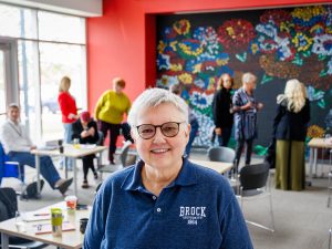 A smiling woman in a Brock University polo shirt poses in a meeting room, with participants and a colourful floral mural in the background.