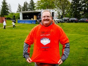 A smiling Rankin Cancer Run participant stands on a rainy grassy field, holding out his bright orange event T-shirt.
