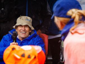 A man in a blue rain jacket smiles while speaking into a microphone during the Rankin Cancer Run opening remarks.