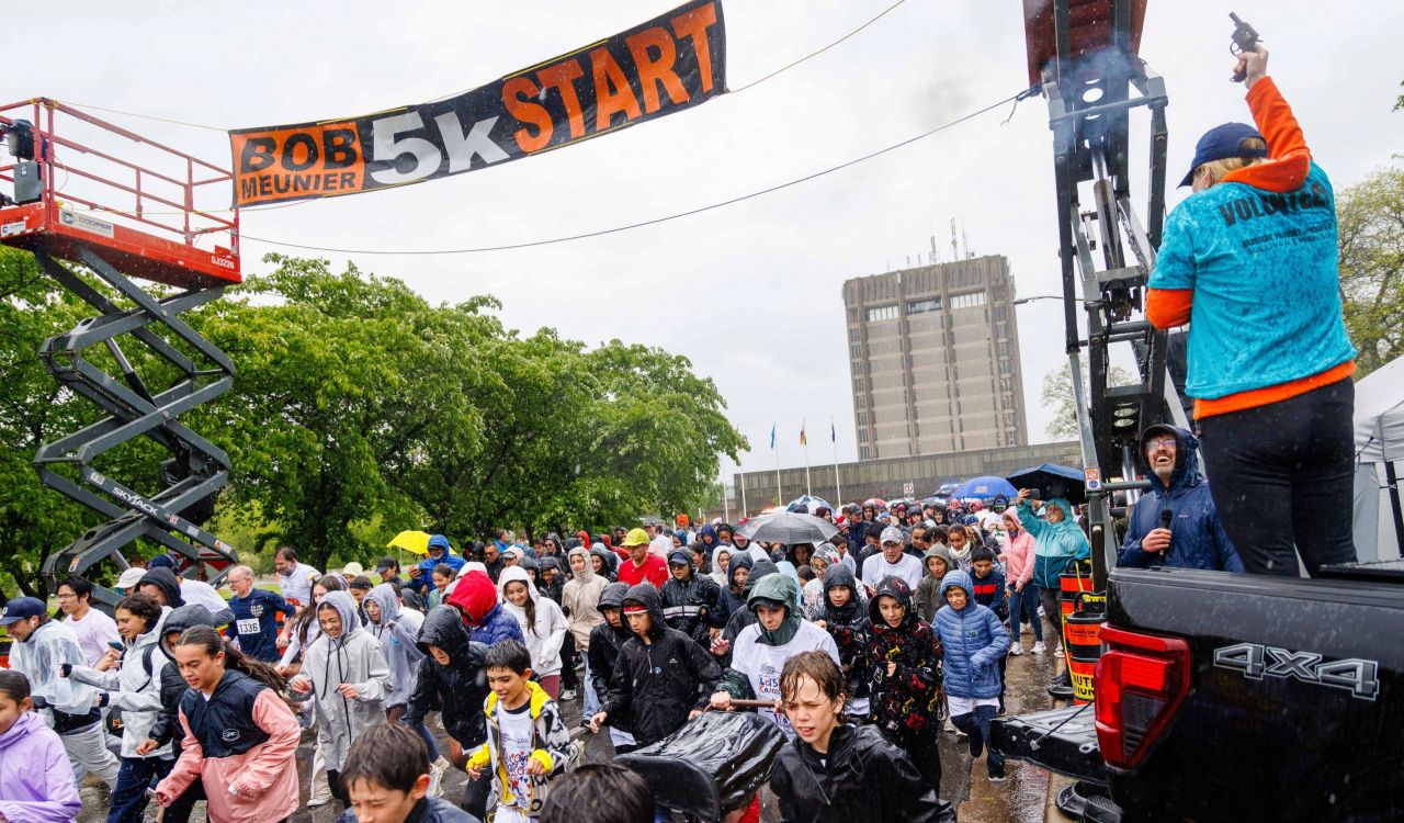 Crowd of children and adults run through heavy rain under the ‘Bob Meunier 5K Start’ banner as volunteers cheer them on from a pickup truck at the Rankin Run.