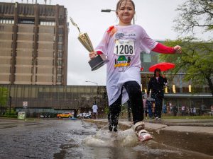 A young Rankin Cancer Run participant carries a trophy while splashing through a puddle in front of Brock University’s Schmon Tower.