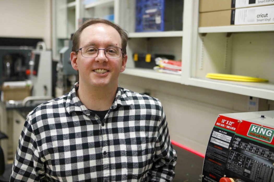 Head-and-shoulders view of a smiling Ryan Schroeder standing in a lab, next to a machine, with equipment fuzzed out in the background.