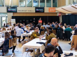 People sitting at tables listen to a woman speak from a podium at an event.