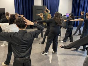 A group of students stand in a studio moving in unison led by a teacher.