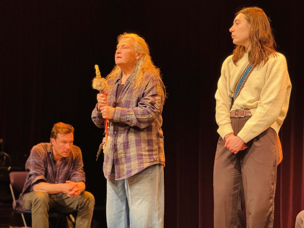 Three performers stand on stage together under stage light.