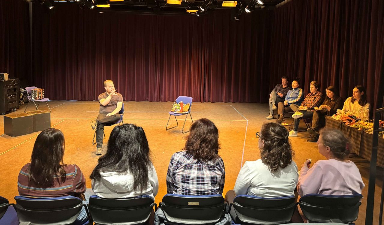 A man sits in a theatre studio speaking to a group of people under stage lights.