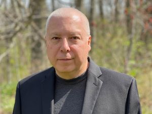 A man wearing a suit poses for a photo outside with trees behind him.