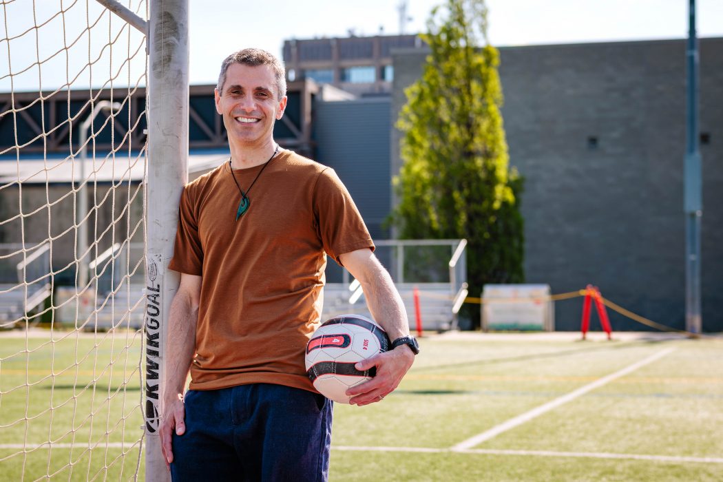 A man poses against a soccer goal post while holding a soccer ball. 