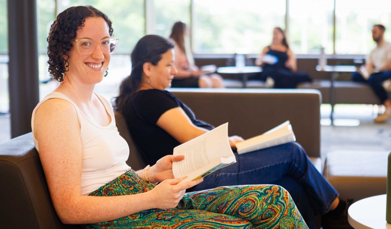 A woman smiles at the camera while holding a book and sitting on a sofa with other people nearby.