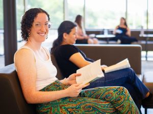 A woman smiles at the camera while holding a book and sitting on a sofa with other people nearby.