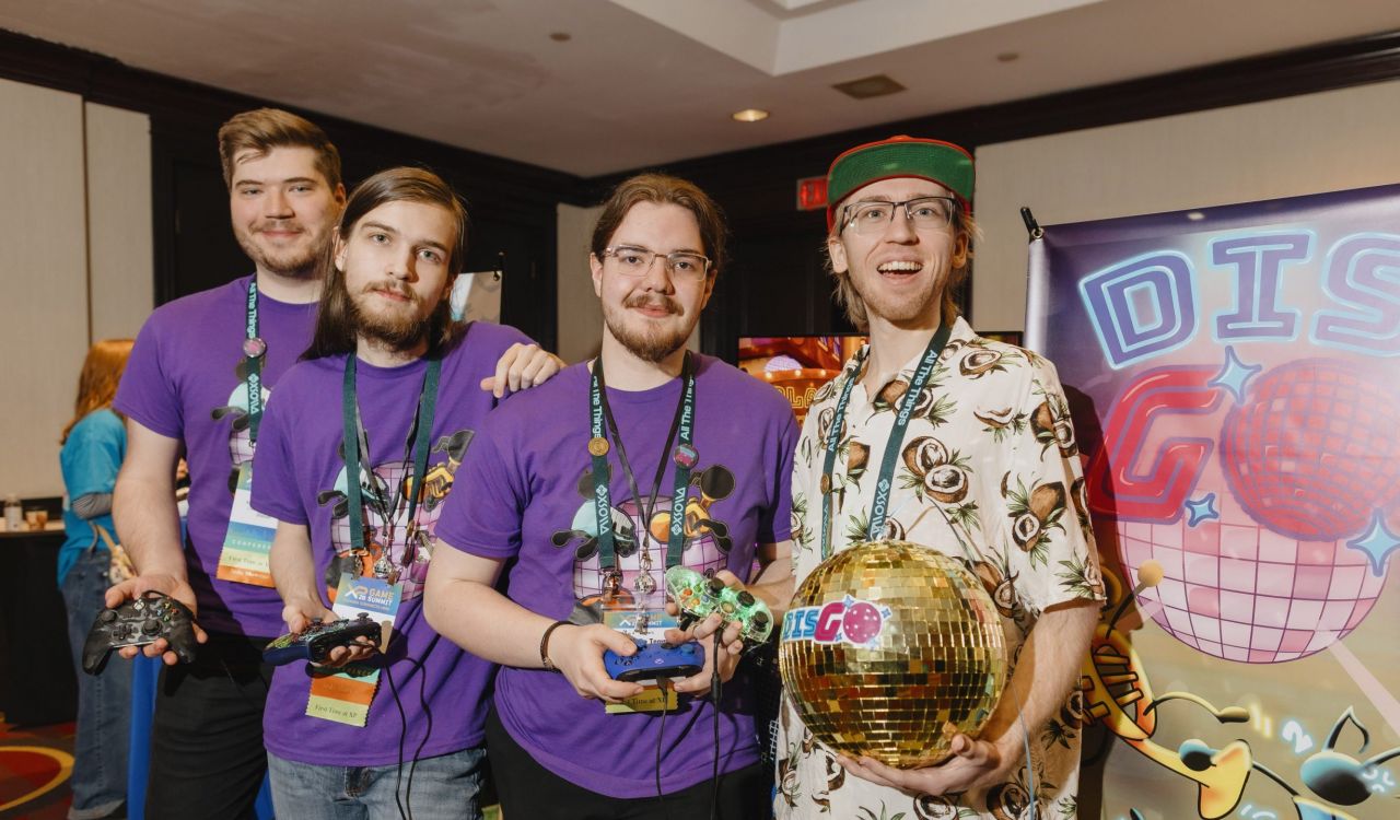 A group of four students hold video game controllers and a disco ball standing together at an event.
