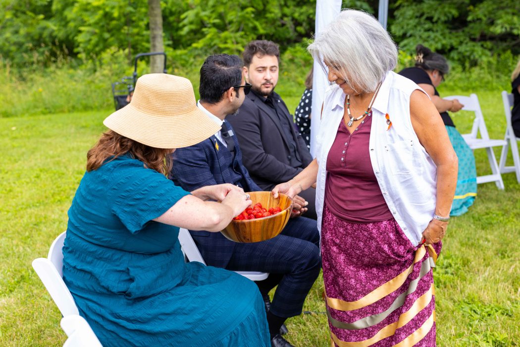 A woman holds a container of strawberries for another woman, seated on a chair, to select one.