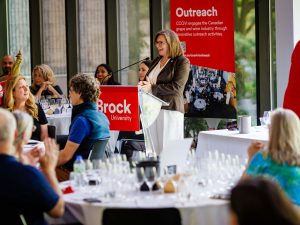 A woman stands at a podium delivering a speech to a group of people seated around round tables.