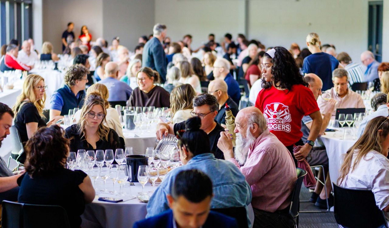 A large group of people sit at round tables tasting flights of wine in a brightly lit room.