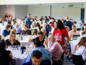 A large group of people sit at round tables tasting flights of wine in a brightly lit room.