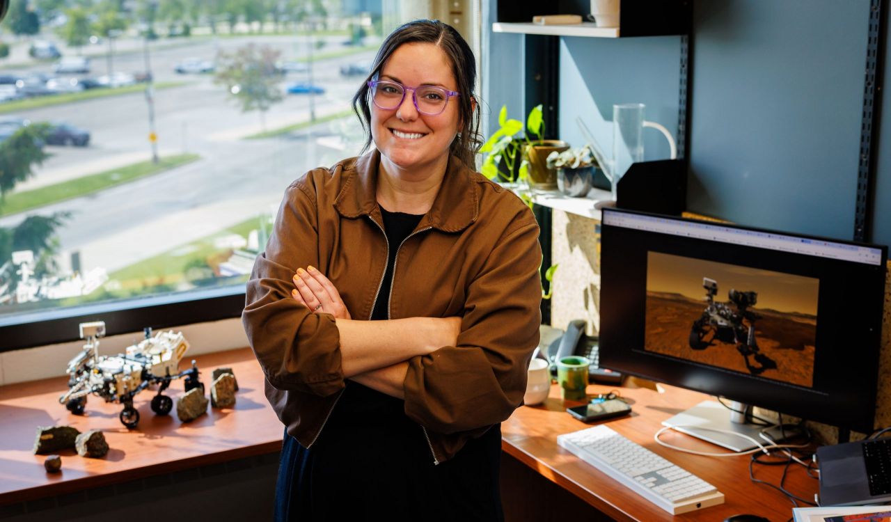 Tanya Kizovski, standing, smiles into the camera, with rocks and a model of the Mars Rover on her left and a computer screen displaying a computer screen with the image of the rover on the right.