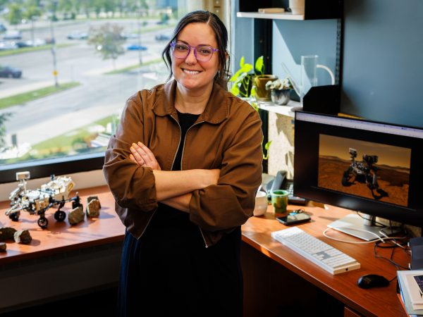 Tanya Kizovski, standing, smiles into the camera, with rocks and a model of the Mars Rover on her left and a computer screen displaying a computer screen with the image of the rover on the right.