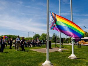 Attendees look up as a Progress Pride flag is raised on a campus flagpole during an outdoor ceremony.