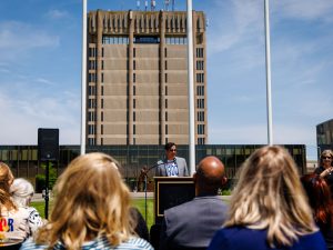 A speaker addresses a seated audience at an outdoor Pride flag-raising ceremony, with Schmon Tower visible behind the podium.