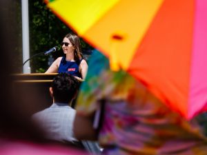 A speaker at a podium smiles while addressing an outdoor audience, partially framed by a brightly coloured Pride umbrella in the foreground.