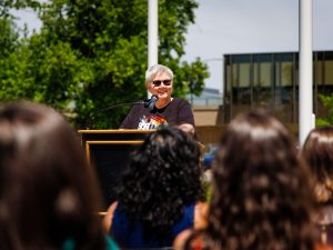 A person in a Brock University Pride shirt speaks at a podium during an outdoor ceremony as audience members watch.