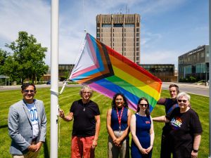 A group poses beneath a raised Progress Pride flag on Brock University’s campus, with Schmon Tower in the background.