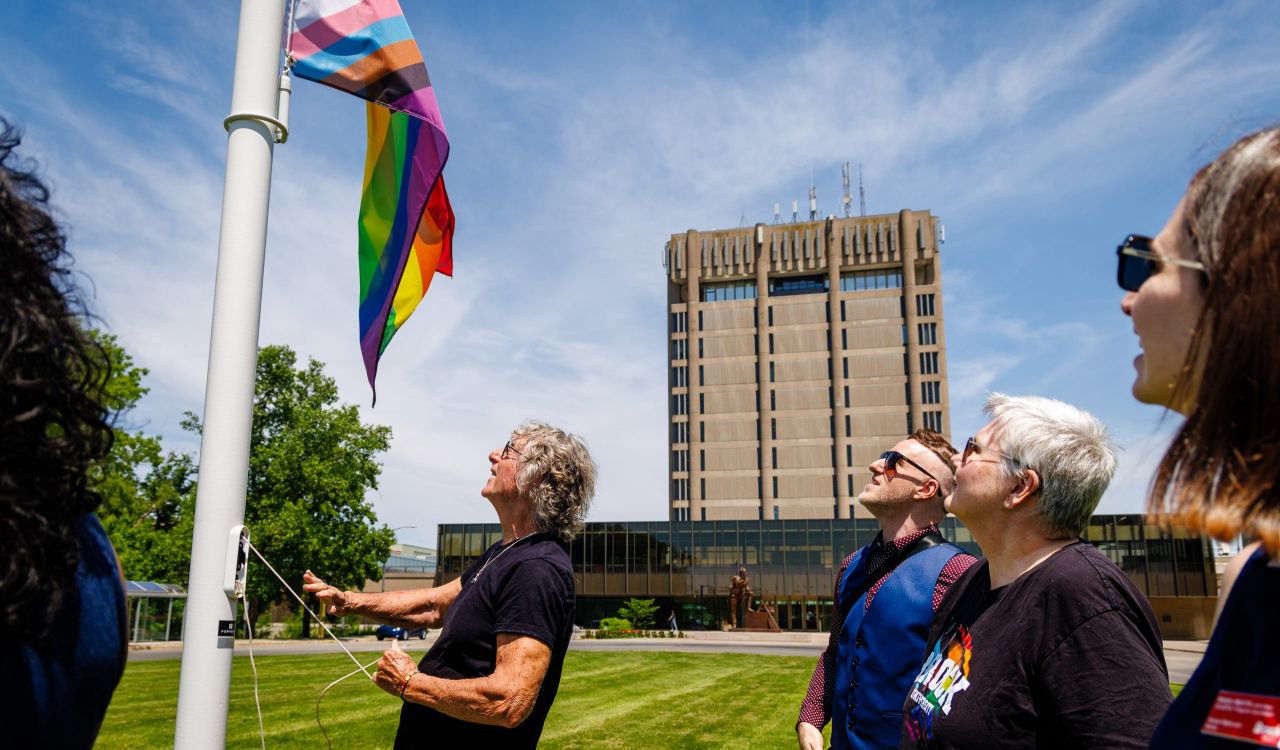 A group of people raise a pride flag outside a tall building.