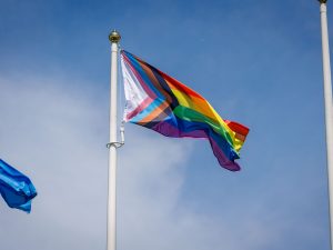 A Progress Pride flag waves high on a flagpole against a blue sky, with another blue flag partially visible nearby.