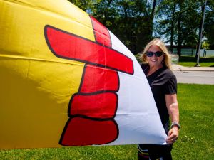 A person stands on a grassy lawn beside a flagpole holding the yellow, red and white Nunavut flag on a sunny day.