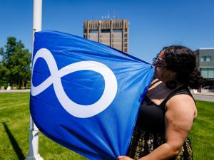 A person stands on a grassy lawn, with the Brock University Schmon Tower in the background, holding the blue and white Métis flag on a sunny day.