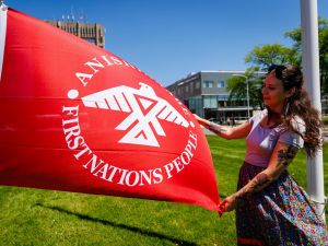 A person stands on a grassy lawn, with Brock University buildings in the background, holding the red and white Thunderbird flag on a sunny day.