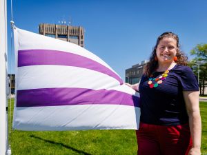A person stands on a grassy lawn, with Brock University buildings in the background, holding the purple and white Two Row flag on a sunny day.