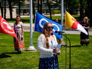A person stands at a microphone outdoors speaking during a flag‑raising ceremony. Three people stand behind them on a lawn holding flags. Trees and campus buildings are visible in the background.