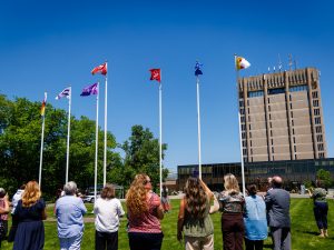 Under a bright blue sky, a group is gathered as Indigenous flags are being raised. In the background is the Brock University tower.