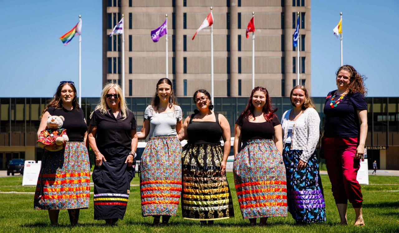 A group of people stand on a grassy lawn with flag poles and University buildings behind them on a sunny day with a clear blue sky.