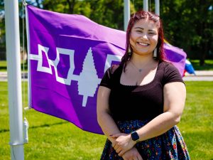 A person stands on a grassy lawn beside a flagpole, with the purple and white Hiawatha flag behind them on a sunny day.