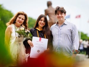 A young woman in an academic gown poses for a photo with loved ones while holding flowers and a diploma.