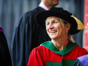 A woman in academic regalia smiles while waiting outside a university graduation ceremony.