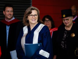 A woman in academic regalia walks ahead of other people in academic regalia before a university graduation ceremony.