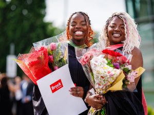 Two smiling young women in academic robes pose for a photo at a university graduation while holding flowers.