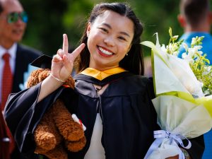 A smiling young woman in academic robes holding flowers and a stuffed toy poses for a photo at a university graduation.