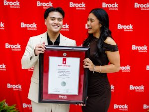 Two people in formal clothing pose holding a diploma at a university graduation celebration.