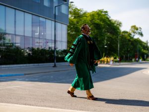 A smiling man in academic robes walks outside after a university graduation ceremony.