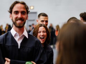 A smiling young woman in academic robes walks stands among classmates at a university graduation.