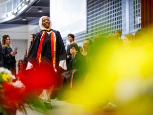 A smiling young woman in academic robes walks across the stage at a university graduation.