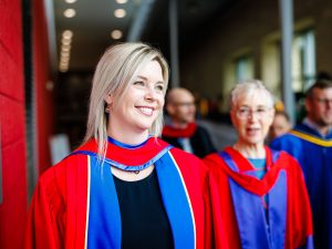 A woman in academic robes smiles outside a university graduation ceremony.